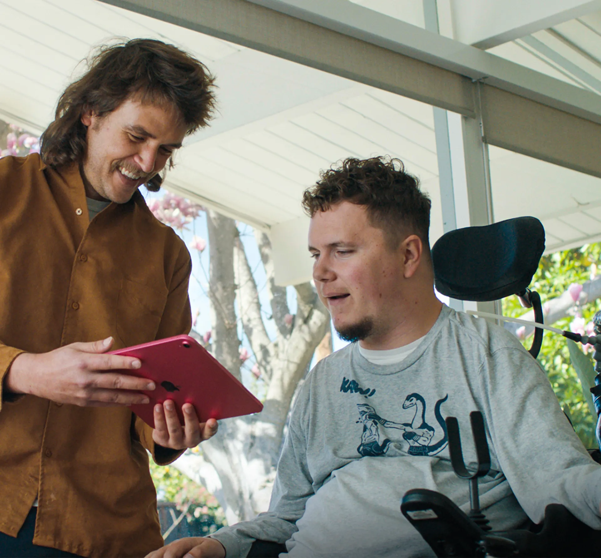 Two friends sharing a laugh over an ipad. one is standing the other in a wheelchair